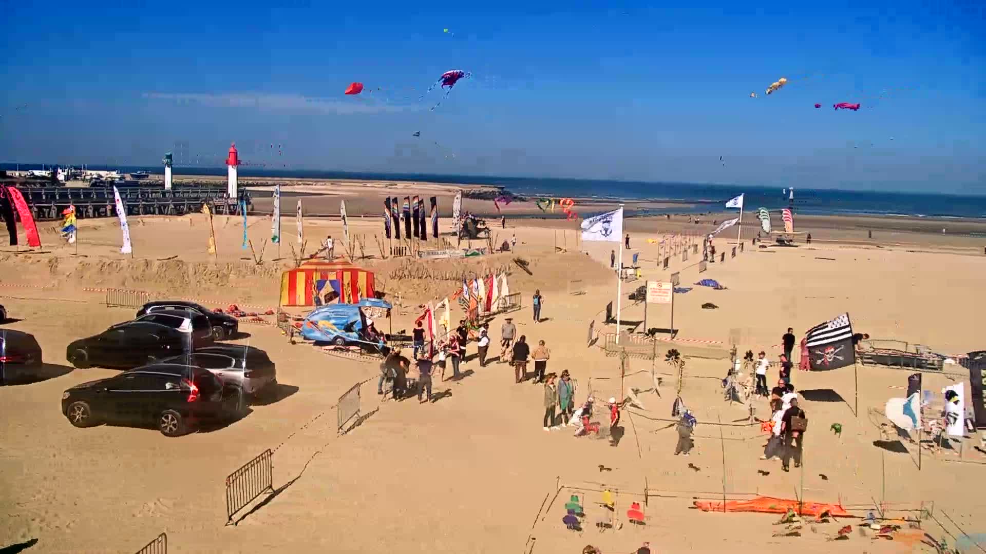 A sandy beach scene on a sunny day shows numerous people gathered around various kites and flags, several cars parked nearby, and a small carnival-like structure.