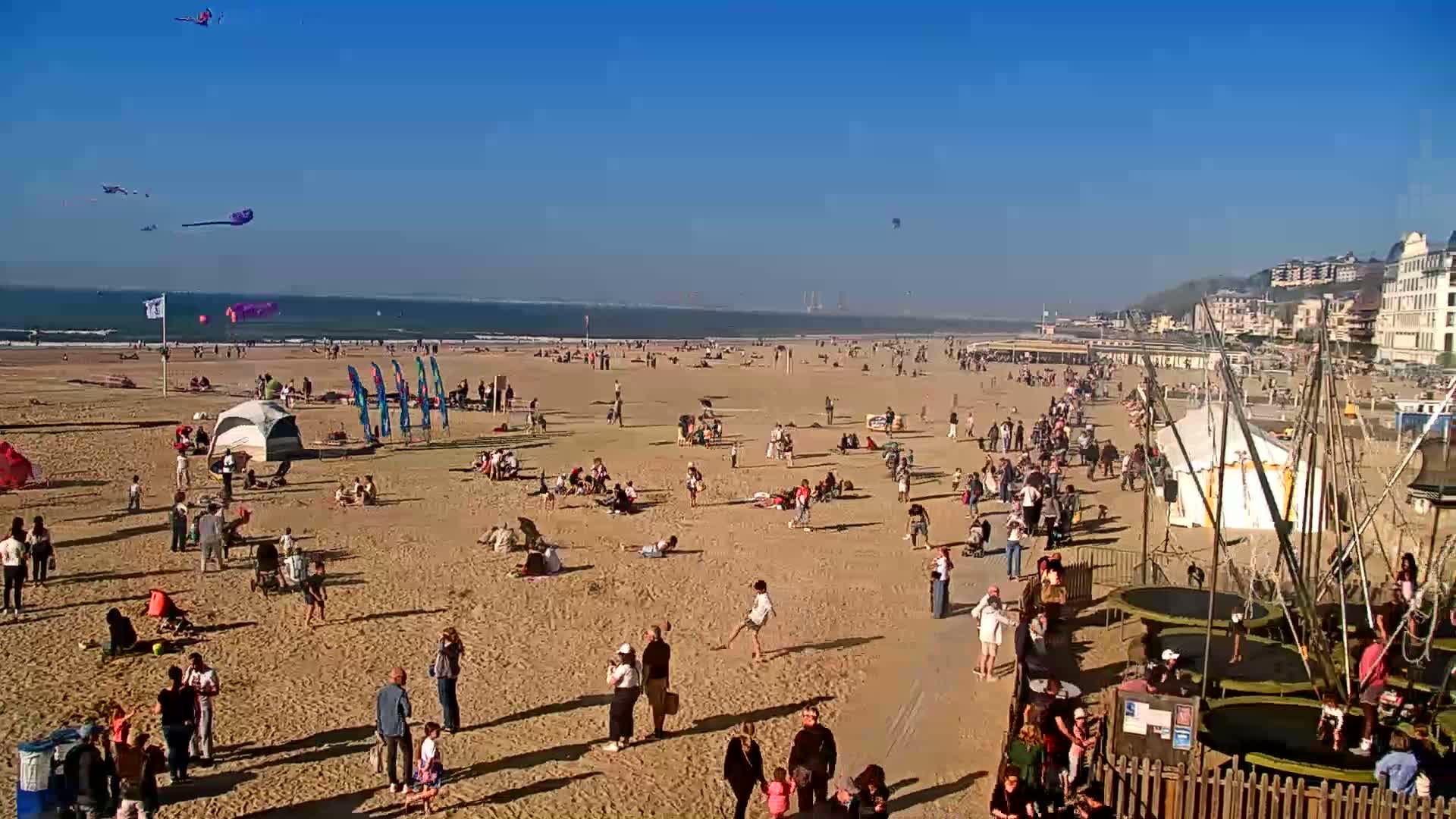 A sandy beach is crowded with people enjoying a sunny day, with several kites flying in the clear blue sky and various beach activities taking place.