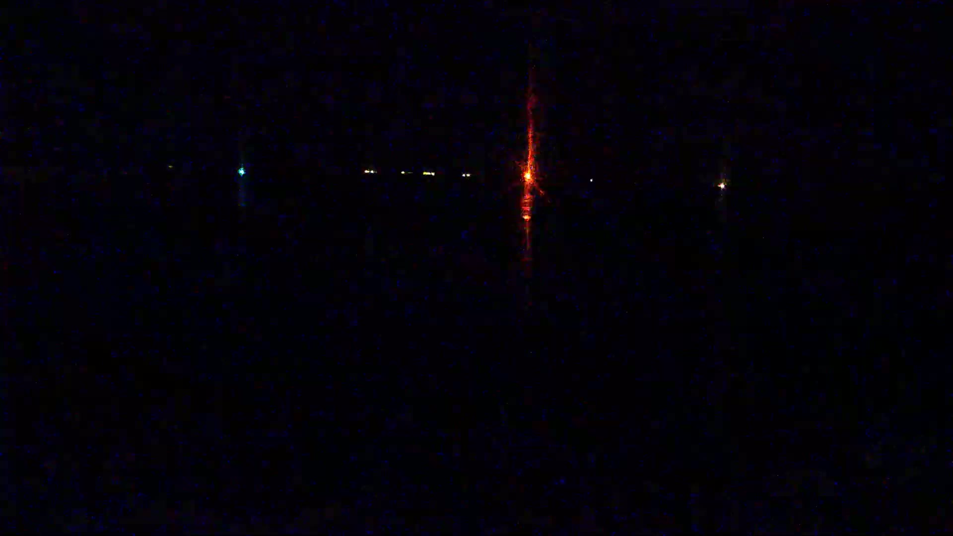A nighttime view of a dark body of water with several small lights and a tall, bright orange light reflecting in the water.