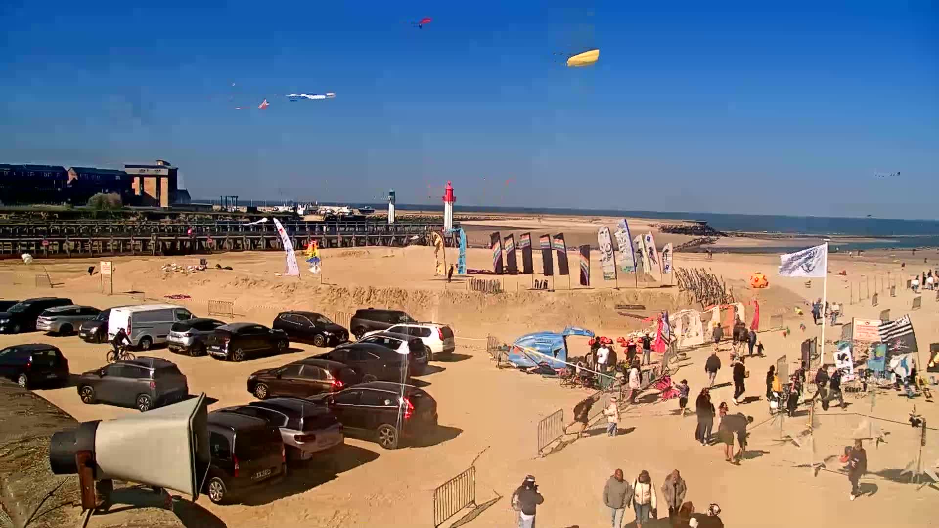 A sandy beach with parked cars, people walking around, several kites in the air, and a few flags and structures on the sand under a clear blue sky.