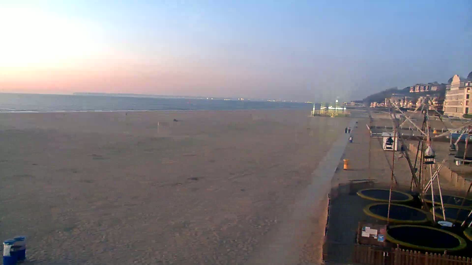 A wide sandy beach at sunset with a calm ocean, a boardwalk, and several trampolines near buildings on the shore.