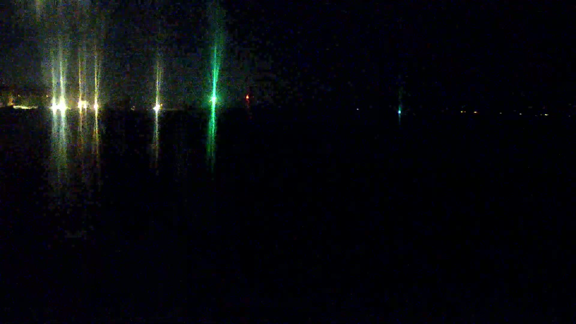 At night, several lights are reflected on a dark body of water, with a few additional lights visible in the distance.