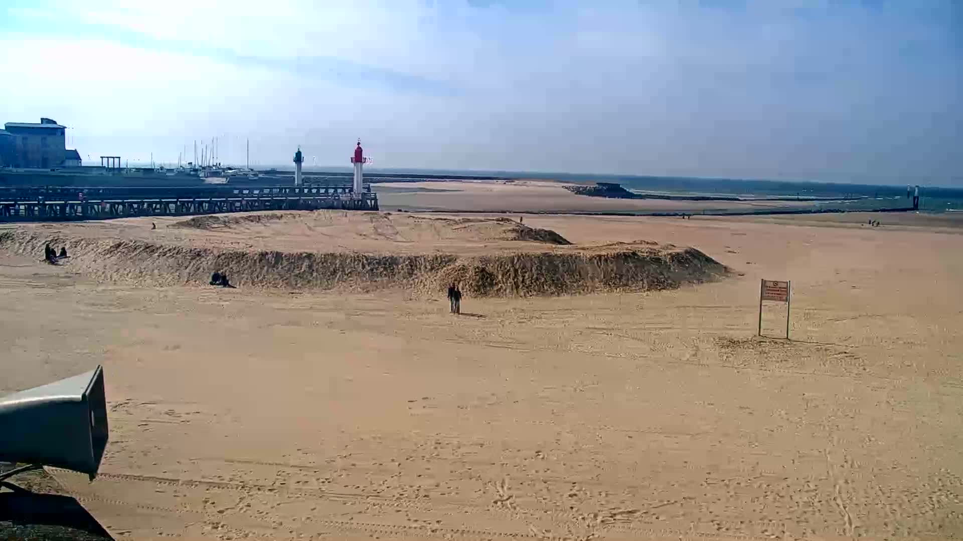 A sandy beach stretches out to a calm ocean under a clear, sunny sky, with a lighthouse and a few people visible in the distance.