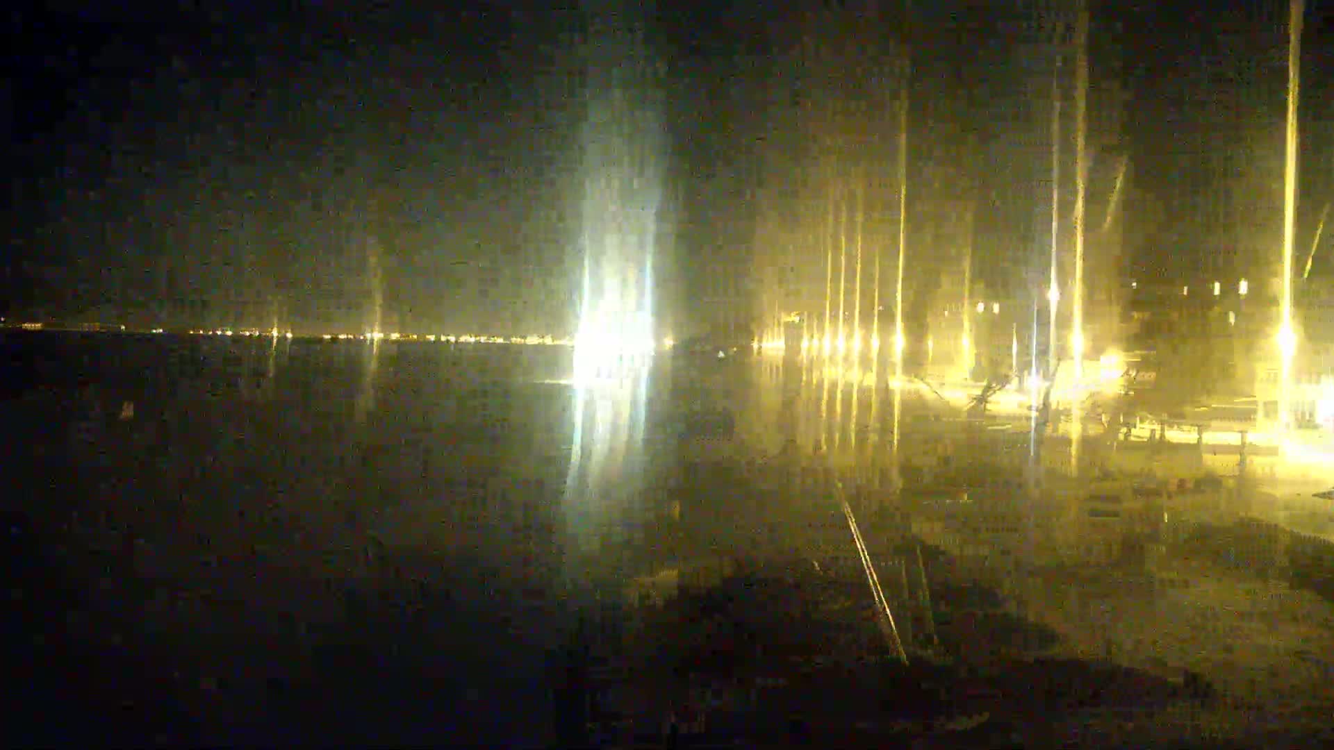 A nighttime view of a calm body of water reflecting the lights of a distant city and a closer, brightly lit waterfront area.