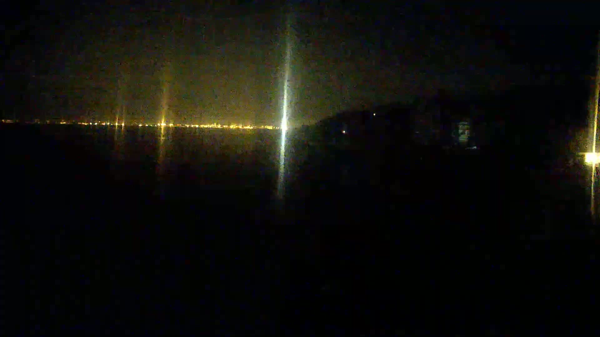 A dark, nighttime scene shows a body of water reflecting the lights of a distant city under a clear sky.