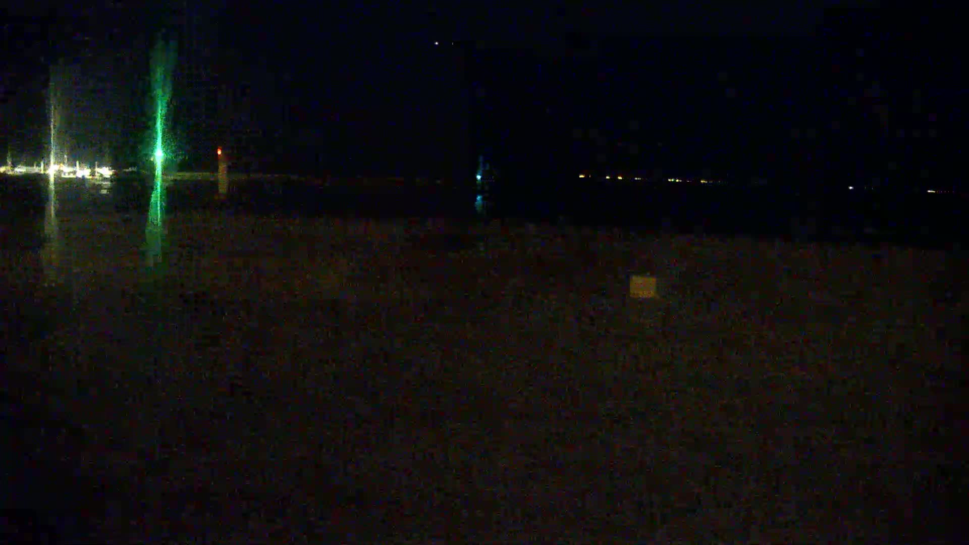 A dark, nighttime scene shows a body of water with faint lights on the opposite shore, a bright green light in the foreground, and a dark, possibly sandy, foreground.
