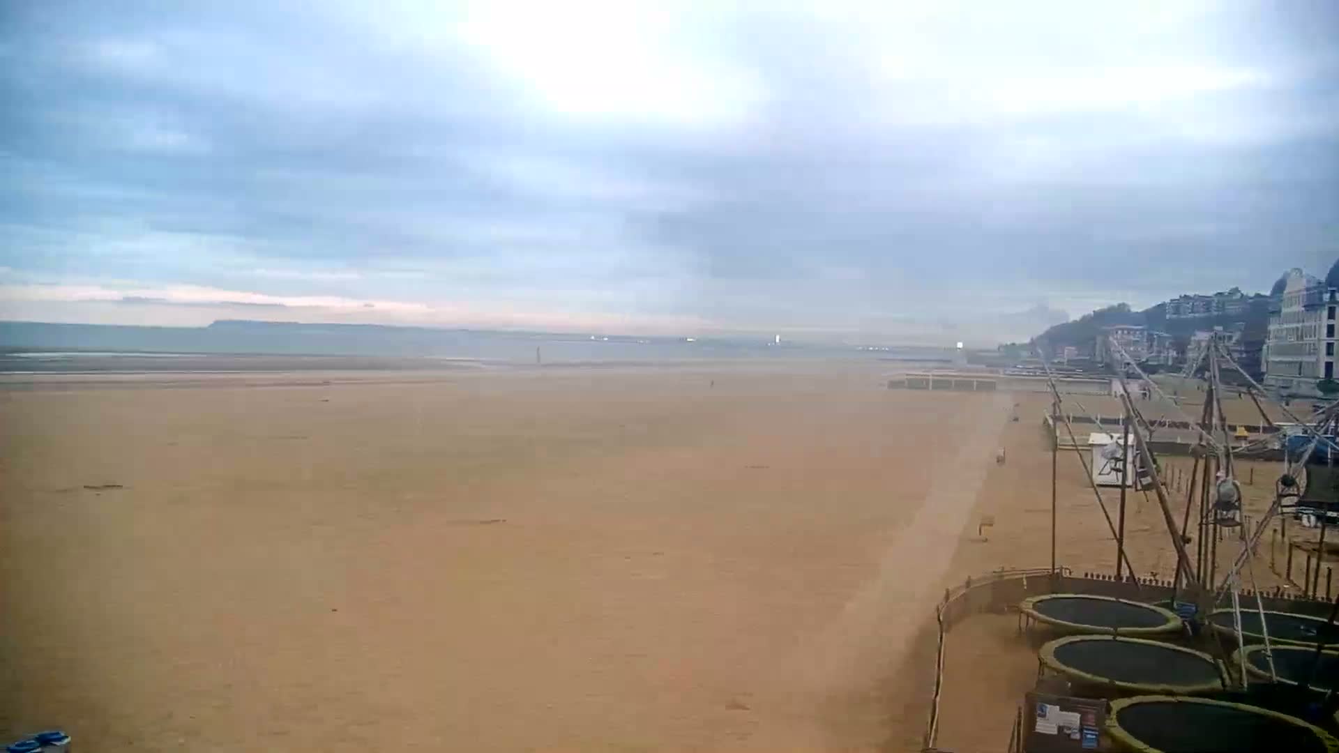 A wide expanse of sandy beach stretches to a calm sea under a cloudy sky, with several trampolines and other beachside structures visible in the foreground.