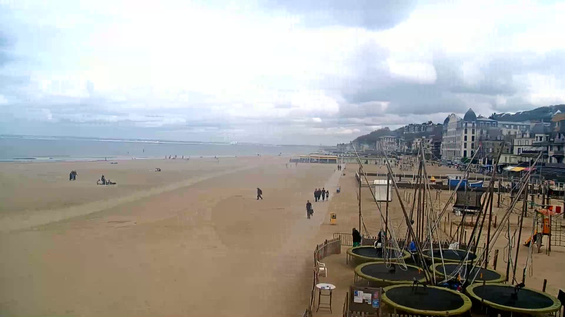 A wide sandy beach with a few people walking and some lying down, several trampolines near the buildings lining the shore, under a cloudy sky.