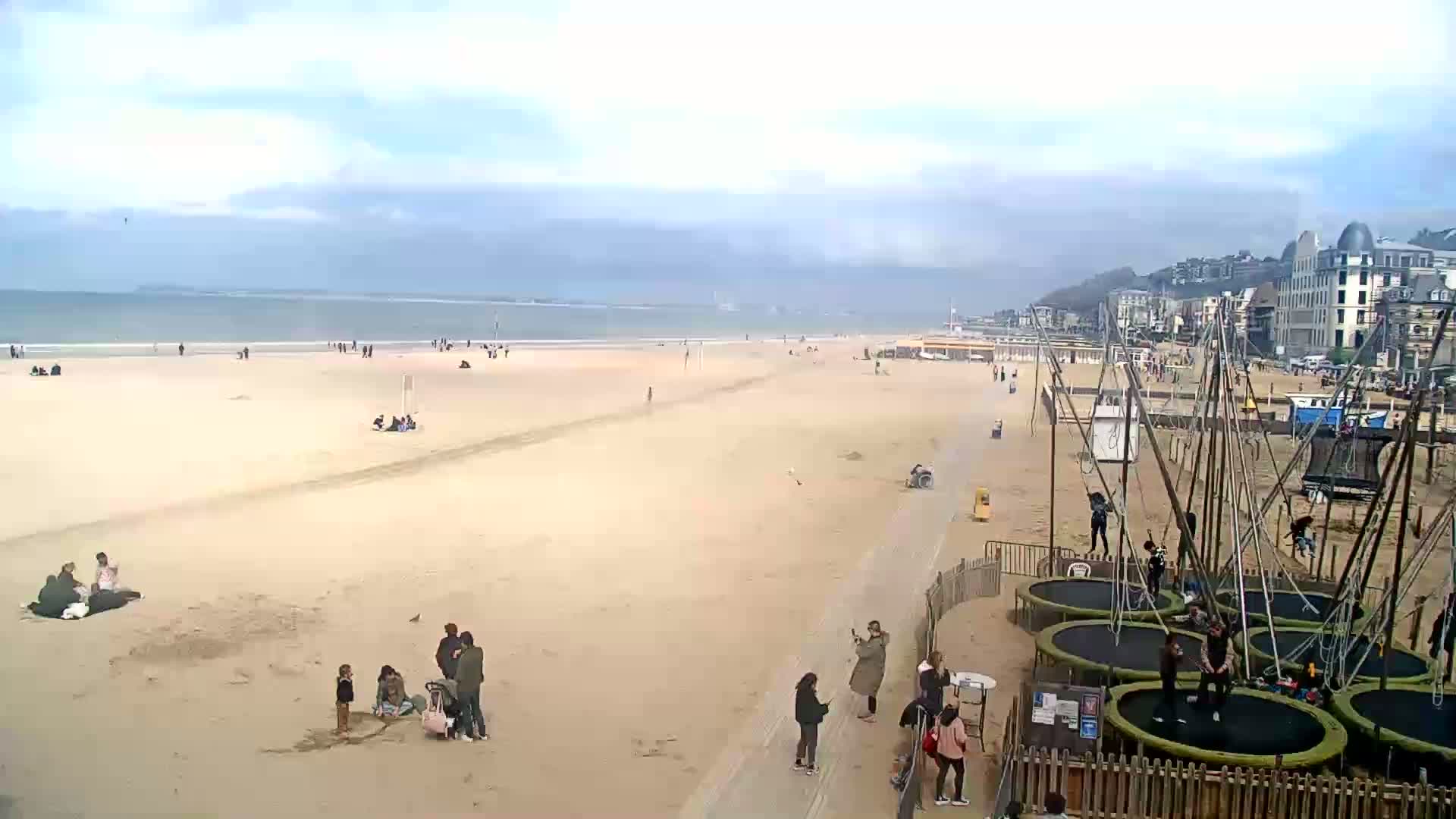 A wide sandy beach with people scattered across it and several trampolines near buildings under a mostly cloudy sky.