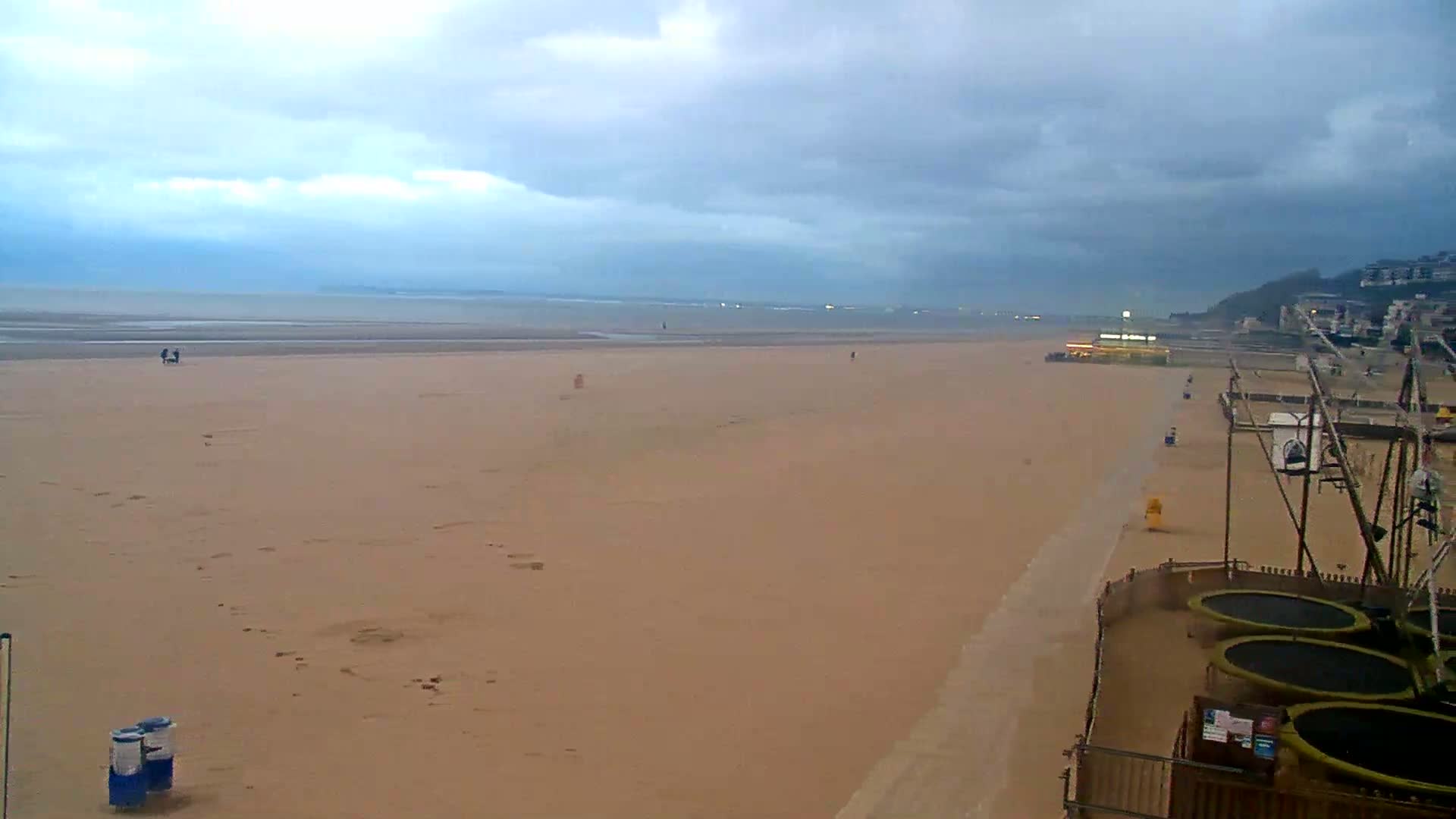 A wide, sandy beach stretches to a calm, overcast ocean under a mostly cloudy sky, with a few people and some beachside amusement equipment visible in the foreground.