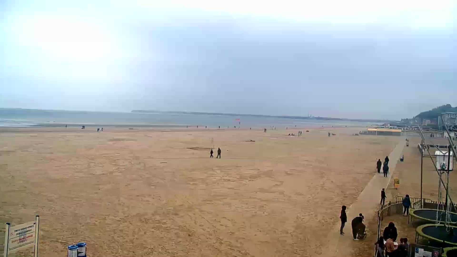 A wide sandy beach stretches to a calm, overcast ocean under a pale sky, with a few people scattered across the sand and near a boardwalk.