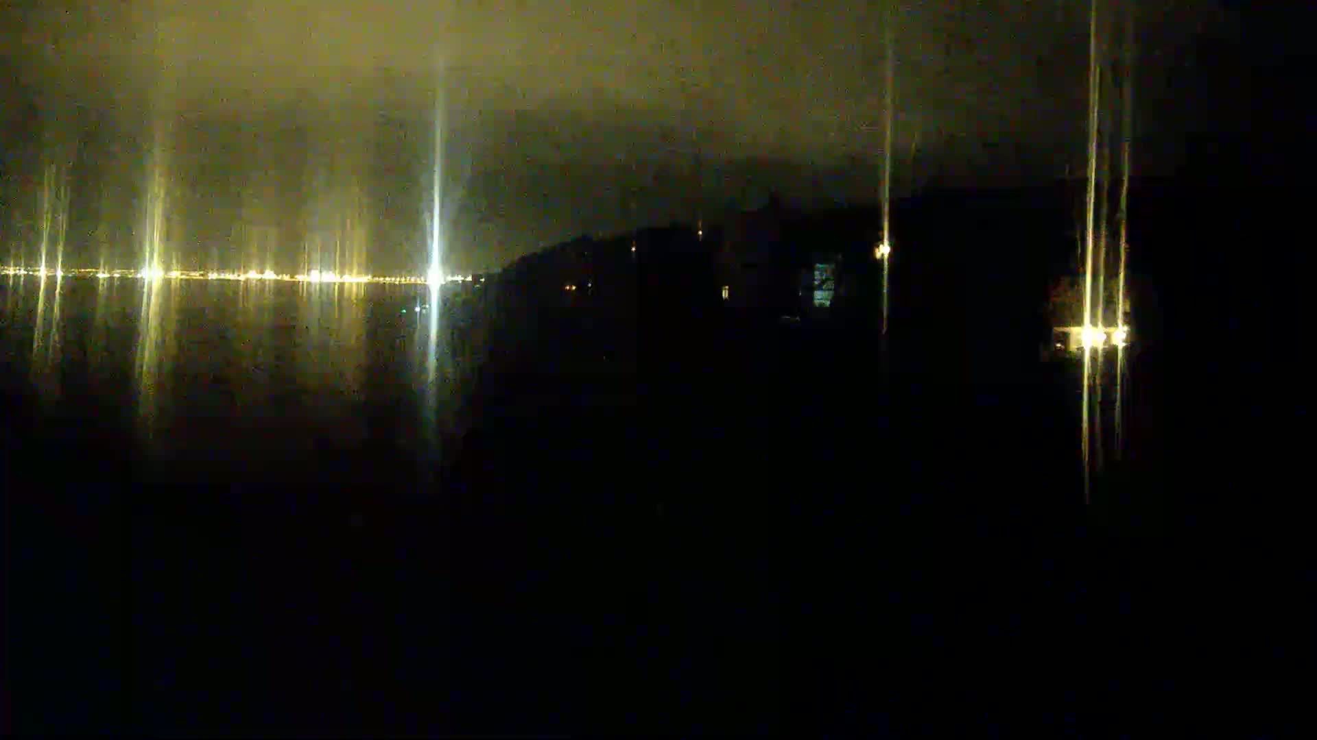 A nighttime view shows a dark body of water reflecting the lights of a distant city under a cloudy sky.
