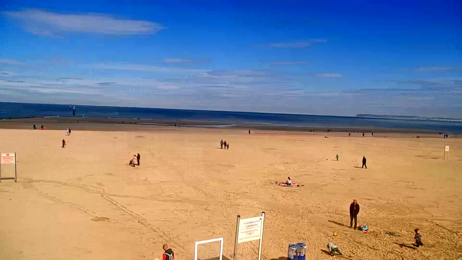 A mostly empty sandy beach stretches to a calm ocean under a clear blue sky with a few scattered clouds.