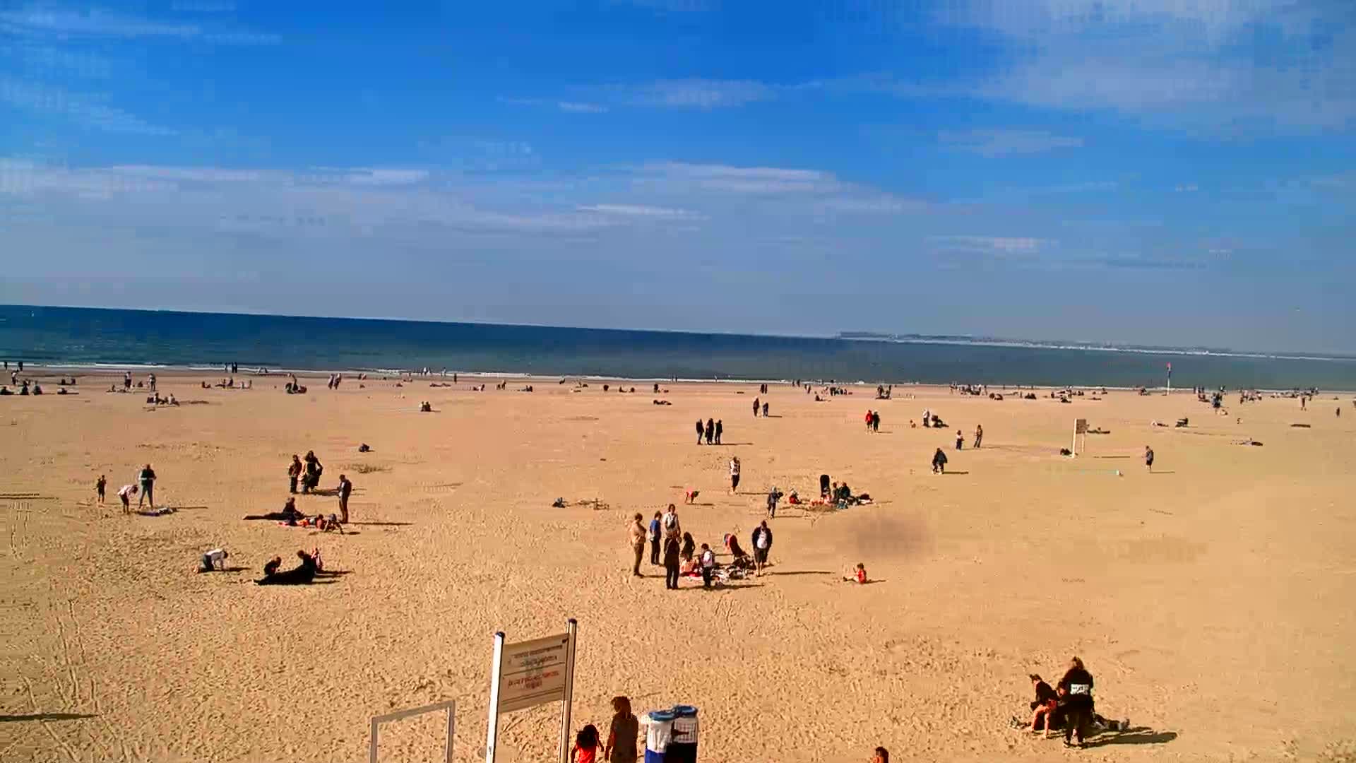 A wide sandy beach with scattered people under a bright sunny sky and a calm ocean in the background.
