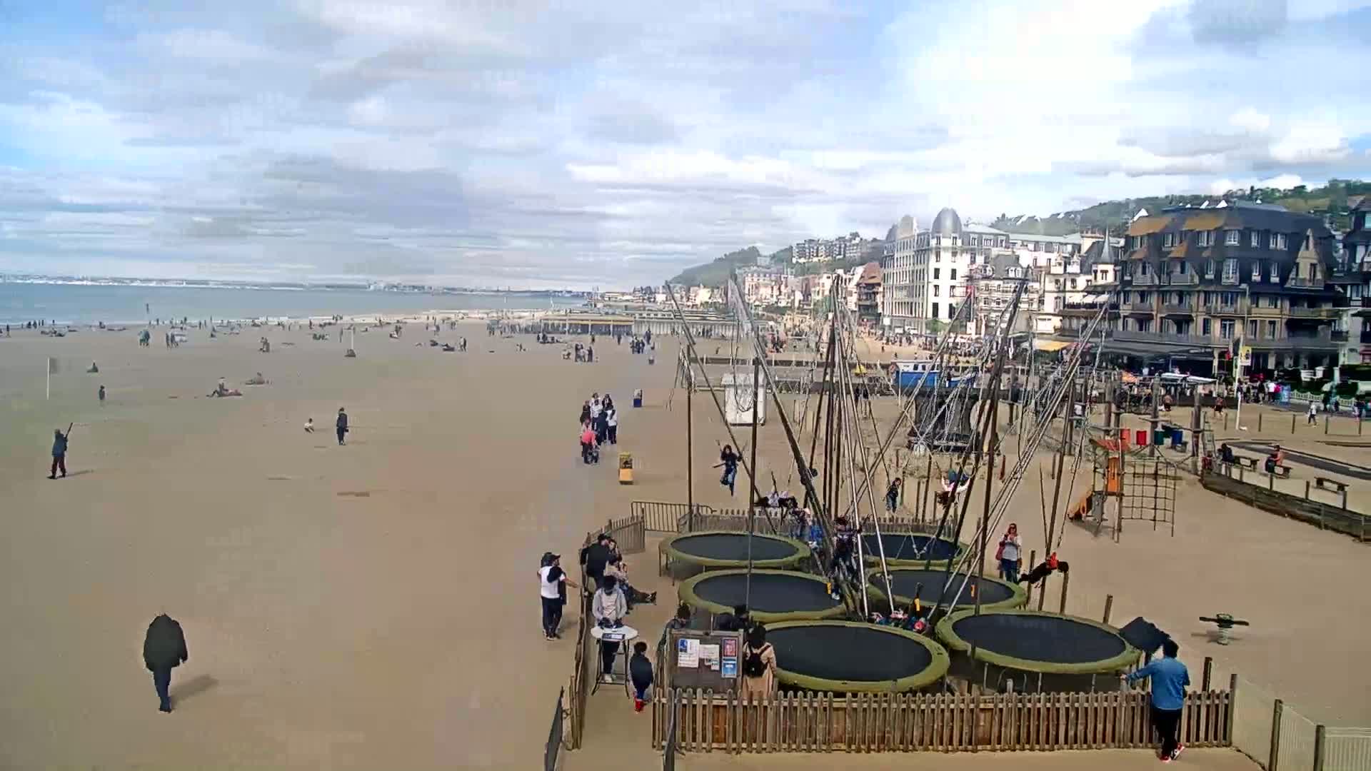 A sandy beach with scattered people, a calm ocean in the background, and several trampolines near buildings under a partly cloudy sky.