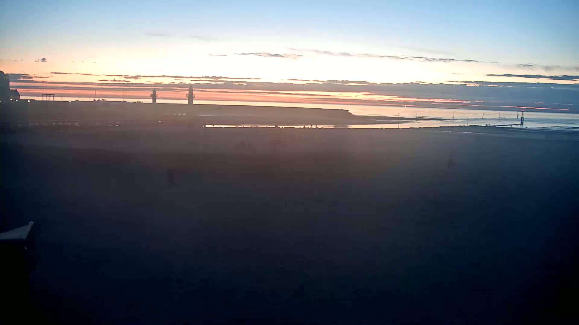 A hazy sunset over a calm body of water, with a beach in the foreground and distant structures on the shoreline.