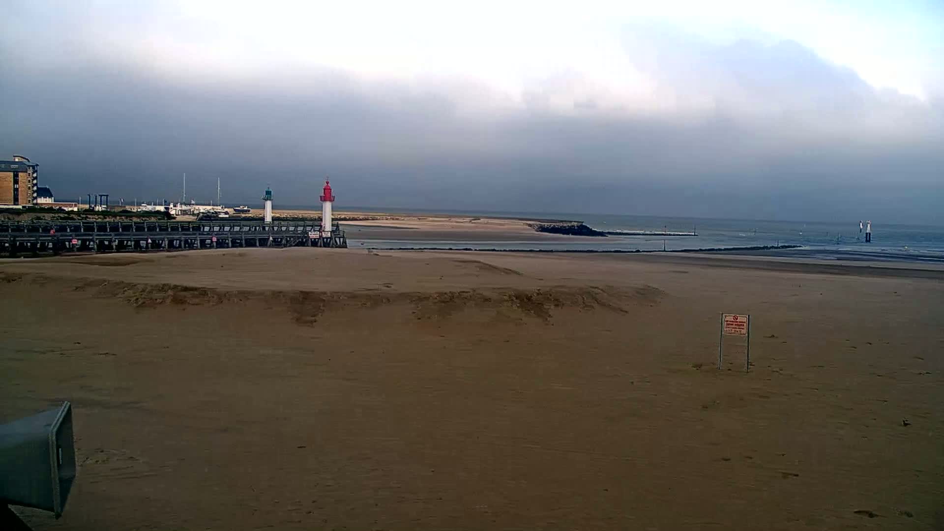 A sandy beach stretches out to a calm sea under a cloudy sky, featuring a pier with two lighthouses and a building in the background.