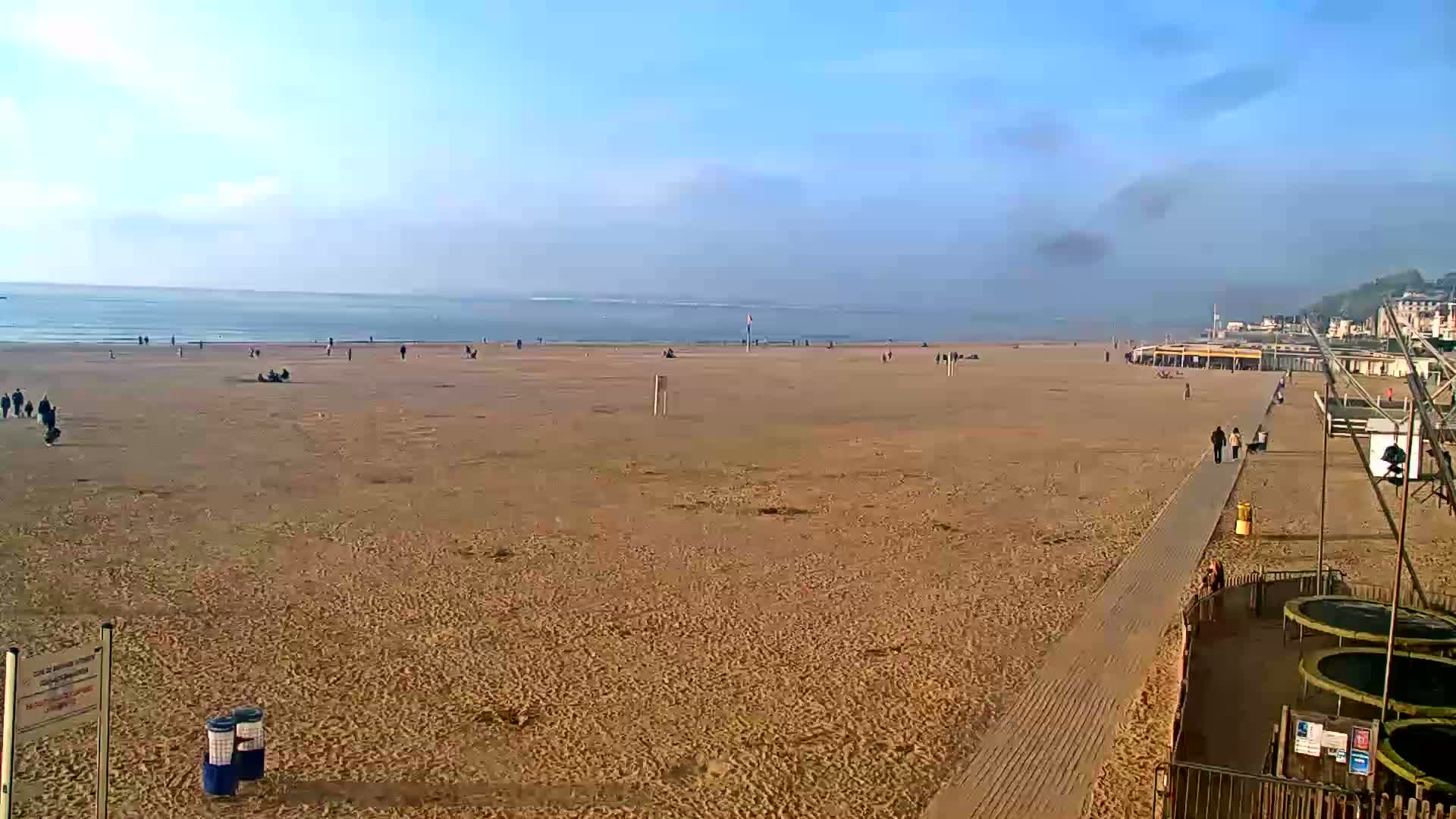 A wide sandy beach stretches to a calm ocean under a clear, bright sky, with a few people scattered across the sand and boardwalk.