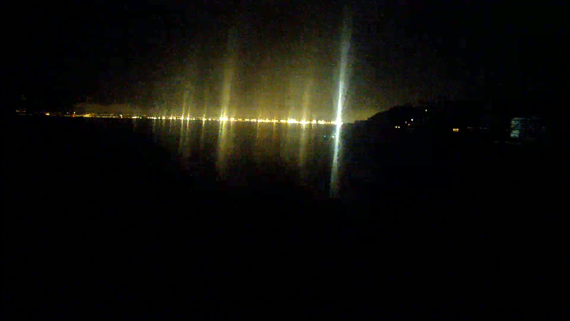 A dark, nighttime scene shows a distant shoreline of lights reflected on calm water.