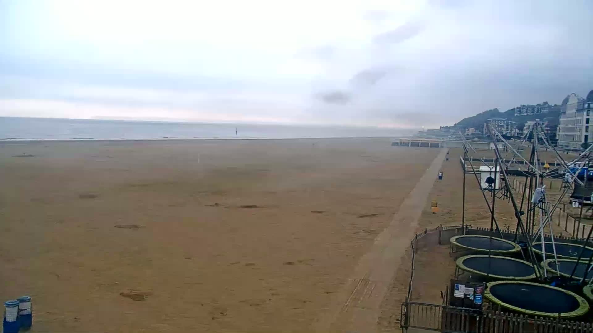 A wide sandy beach stretches to a calm, overcast sea under a mostly cloudy sky, with several trampolines and buildings visible in the distance.