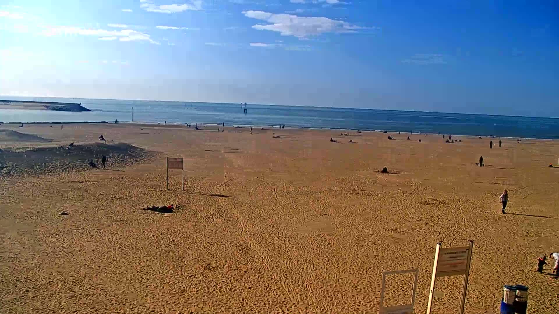 A wide sandy beach with scattered people stretches to a calm ocean under a mostly clear, sunny sky.