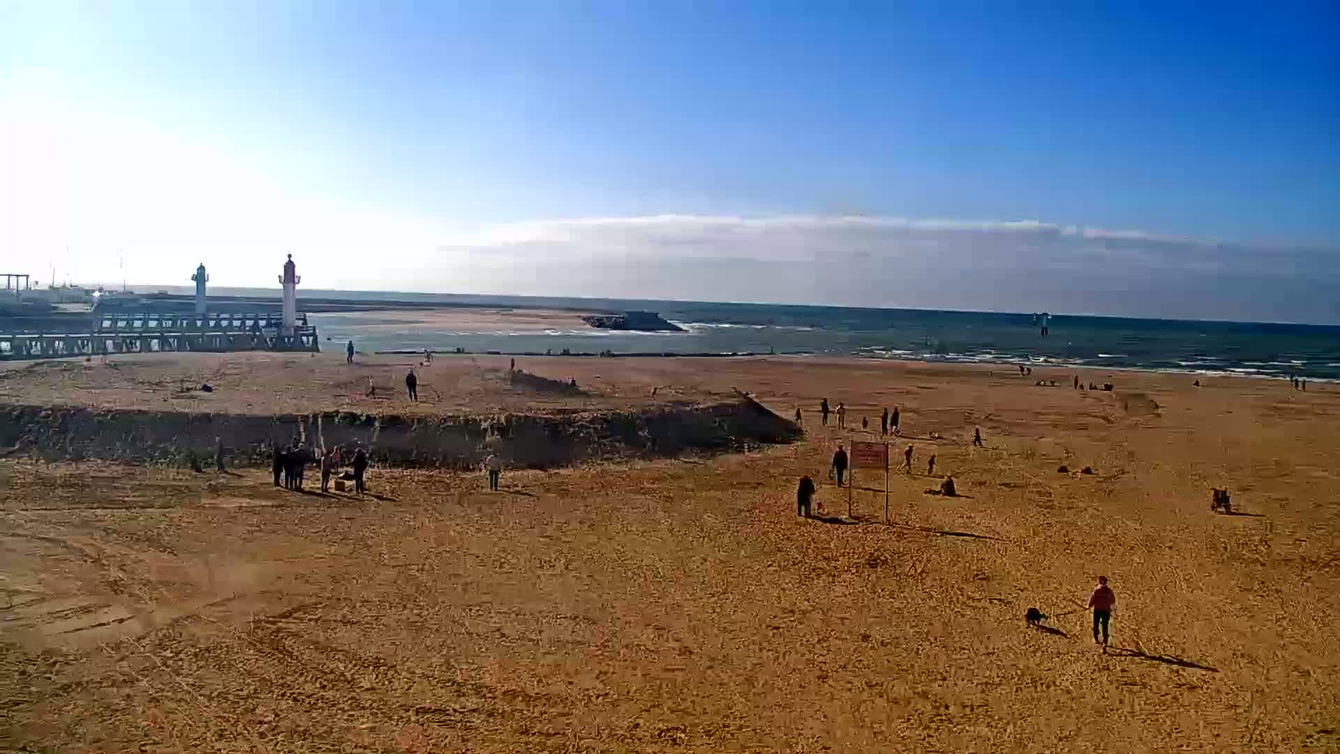 A sandy beach with scattered people and a few dogs, a pier with two lighthouses, and a calm ocean under a mostly sunny sky.