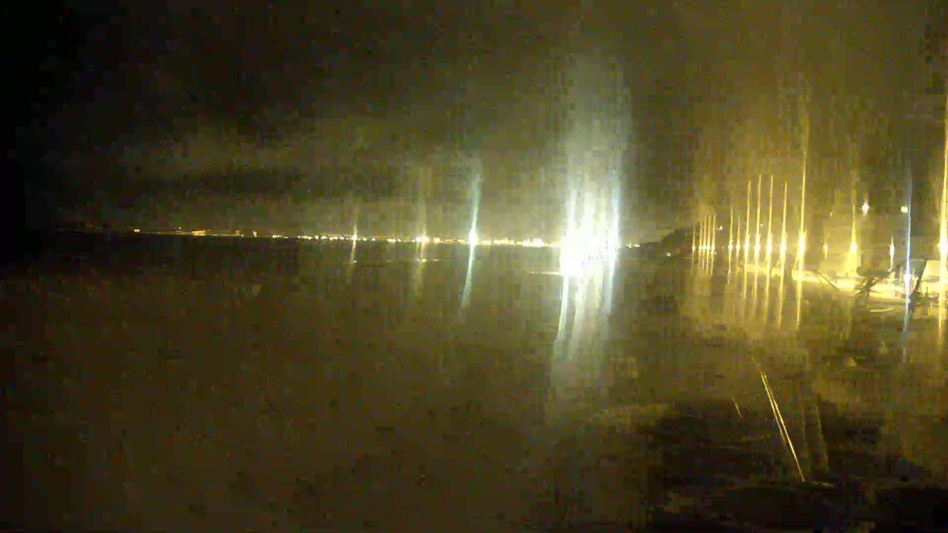 A nighttime view of a body of water reflecting the lights of a distant city and a closer shoreline with illuminated poles.