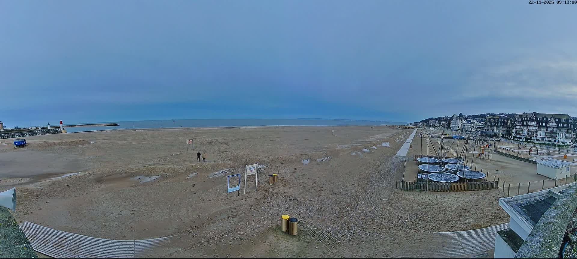 A wide, overcast beach stretches under a grey sky, featuring a pier with a lighthouse on the left, two individuals walking on the sandy shore, and a promenade lined with buildings and covered trampolines on the right.