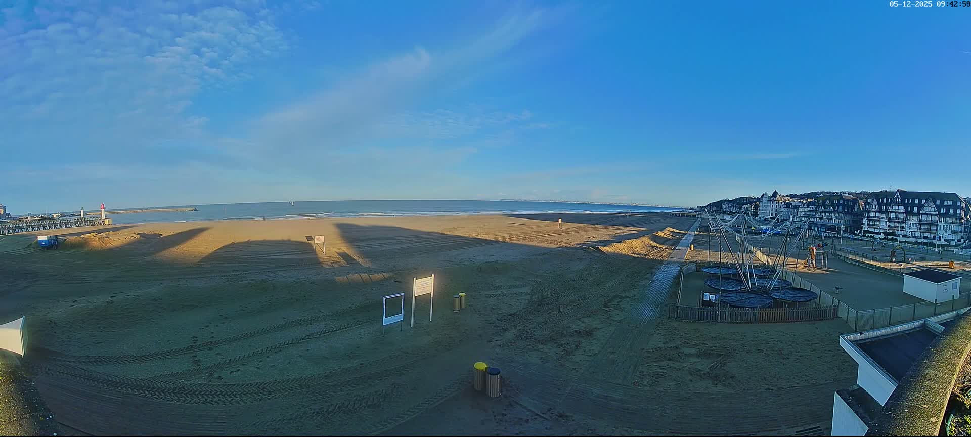 A wide sandy beach with visible tire tracks and long shadows stretches along a coastline featuring a pier with a lighthouse, distant ships on the calm sea, and a town built into a hill, all under clear blue skies with scattered white clouds.
