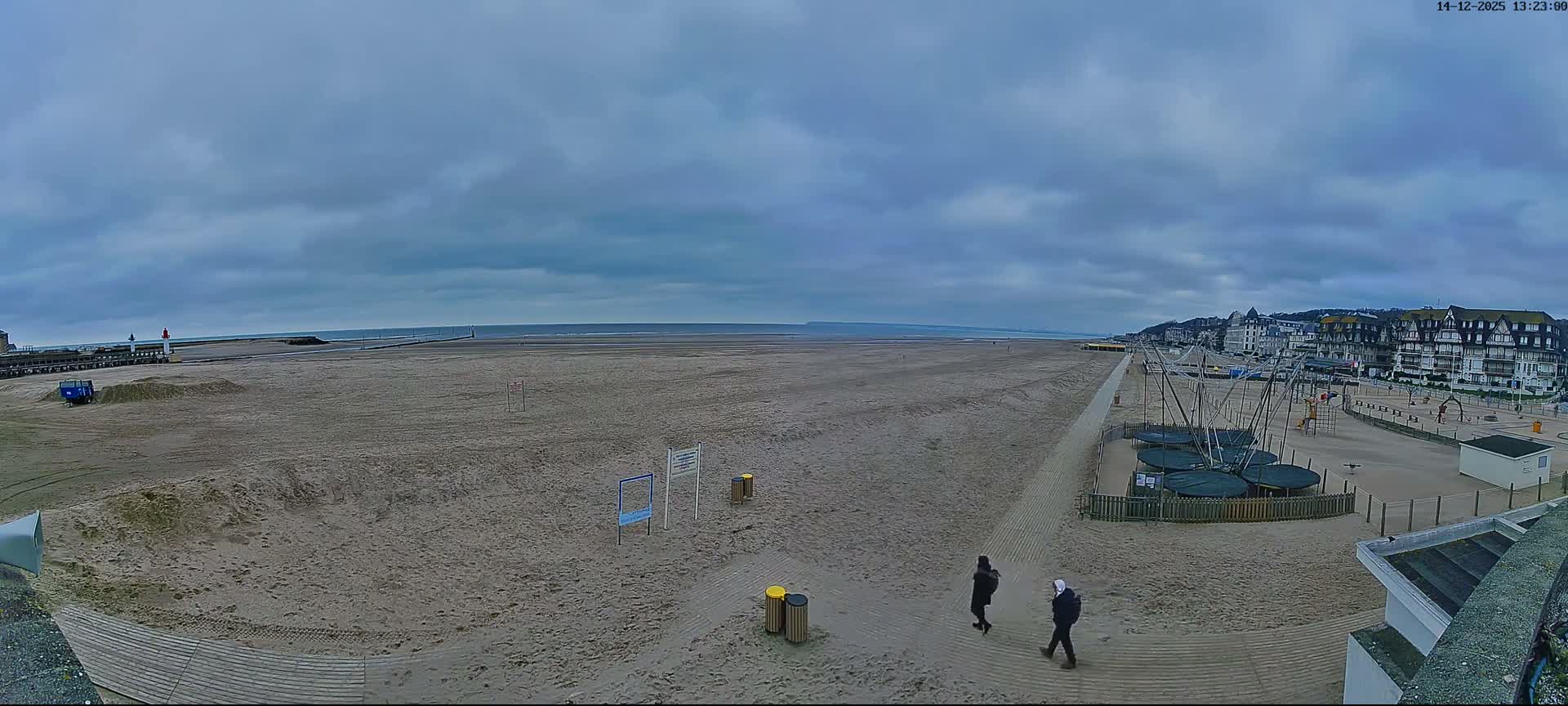 A wide sandy beach with visible tire tracks and long shadows stretches along a coastline featuring a pier with a lighthouse, distant ships on the calm sea, and a town built into a hill, all under clear blue skies with scattered white clouds.
