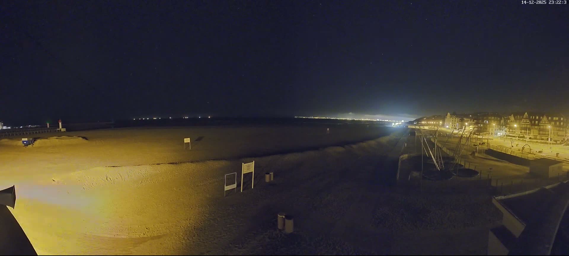 A wide sandy beach with visible tire tracks and long shadows stretches along a coastline featuring a pier with a lighthouse, distant ships on the calm sea, and a town built into a hill, all under clear blue skies with scattered white clouds.