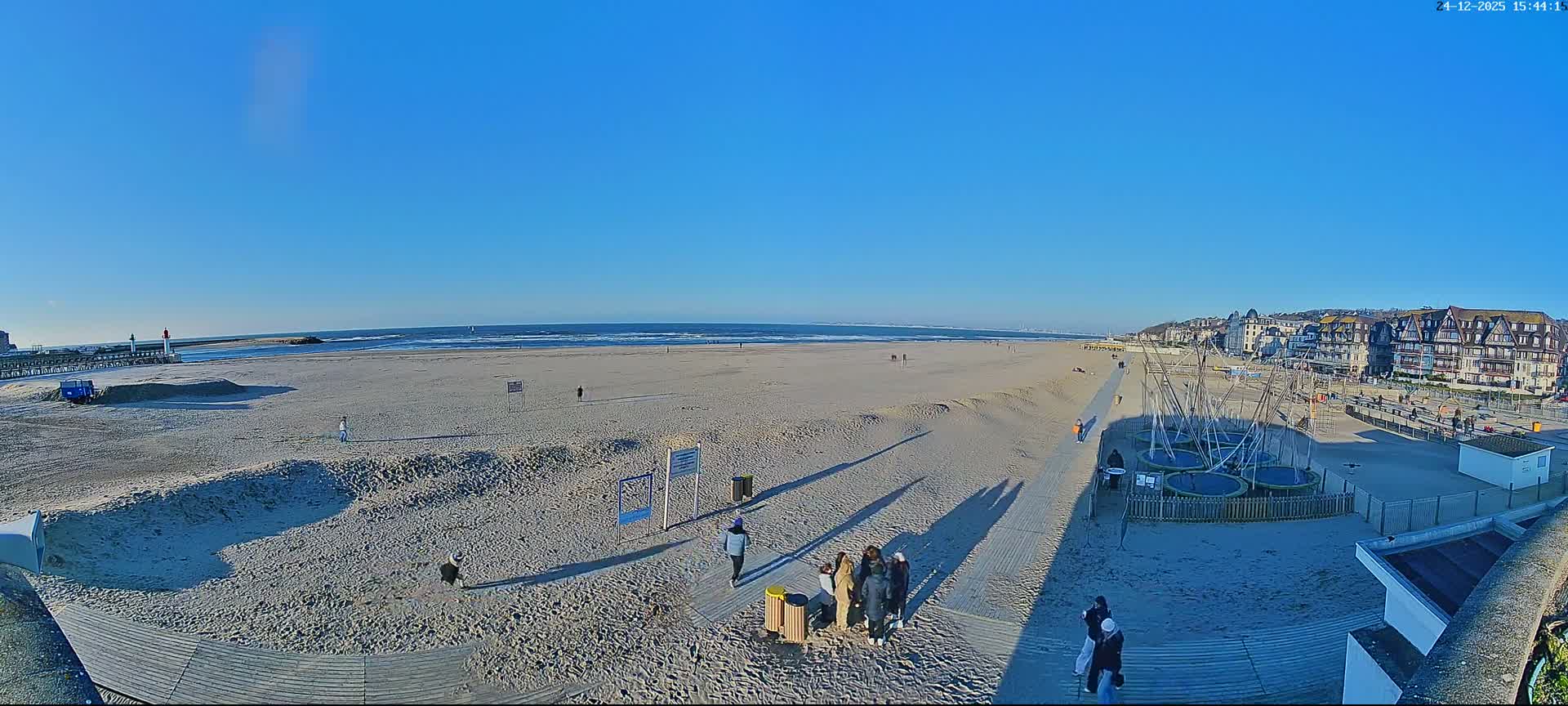 A wide sandy beach with visible tire tracks and long shadows stretches along a coastline featuring a pier with a lighthouse, distant ships on the calm sea, and a town built into a hill, all under clear blue skies with scattered white clouds.
