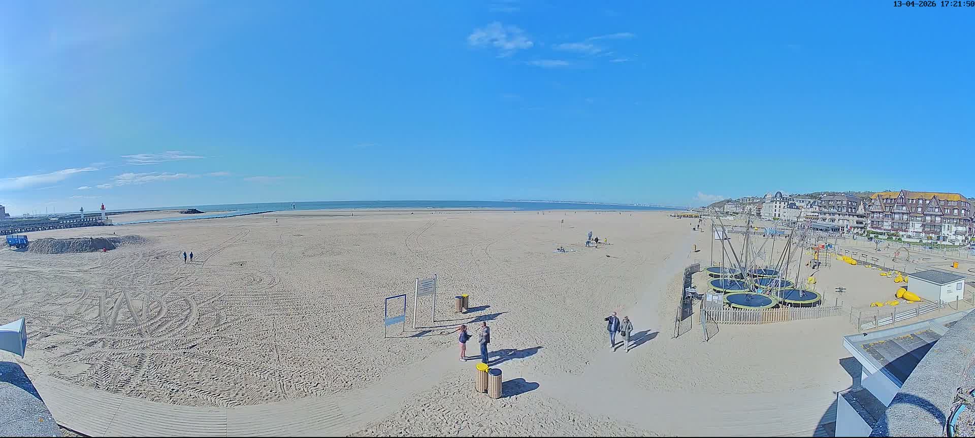 A wide sandy beach with visible tire tracks and long shadows stretches along a coastline featuring a pier with a lighthouse, distant ships on the calm sea, and a town built into a hill, all under clear blue skies with scattered white clouds.