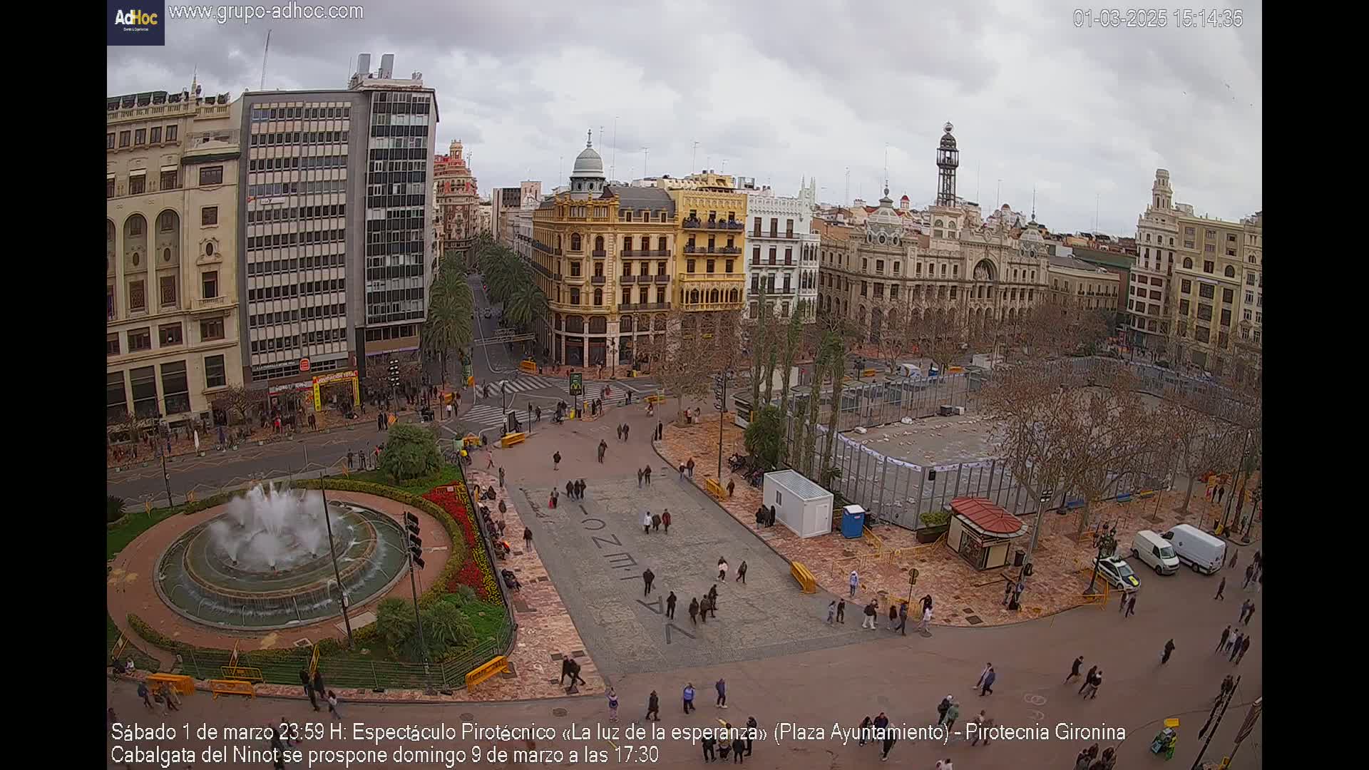 A city square with a fountain, several buildings, and many people is seen on an overcast day.
