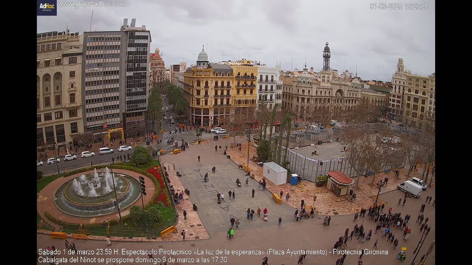 A city square with a fountain, surrounded by buildings, with many people gathered under an overcast sky.