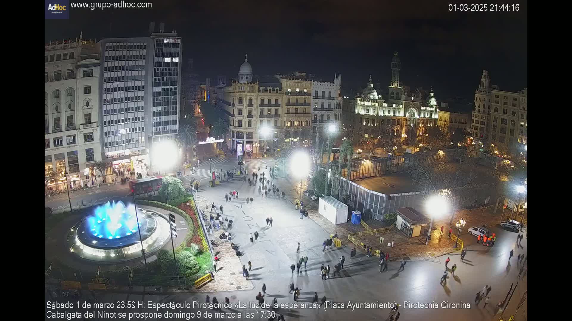A nighttime aerial view of a city square with a fountain, many people walking around, and buildings lit up.