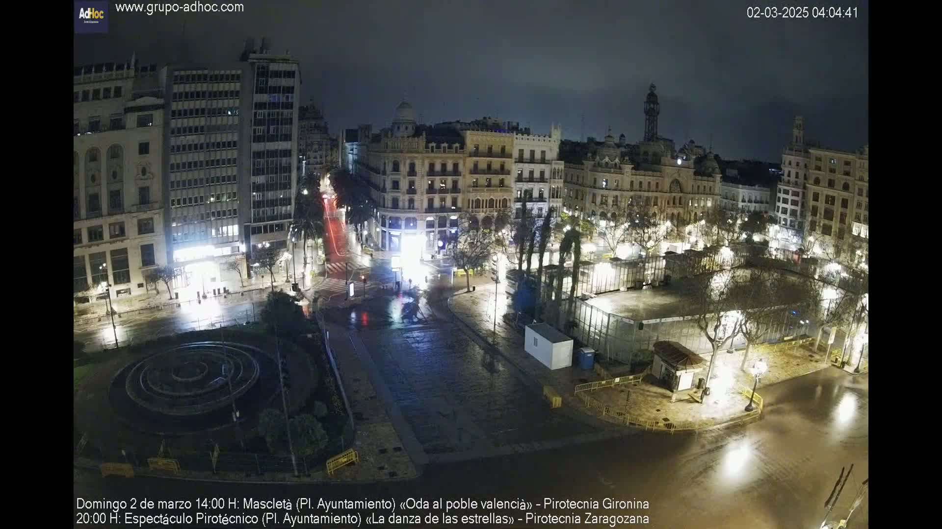 A nighttime view of a city square with wet streets, illuminated buildings, and sparsely populated areas under a dark sky.