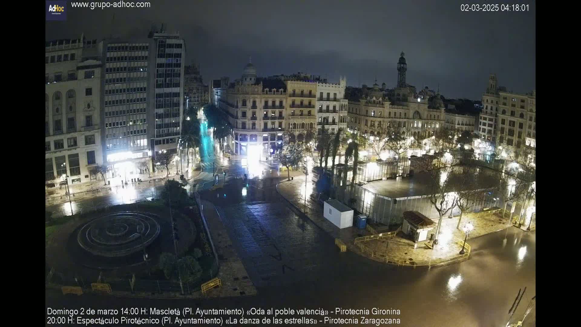 A nighttime, rain-soaked city square with buildings illuminated, a fountain in the center, and temporary structures set up.