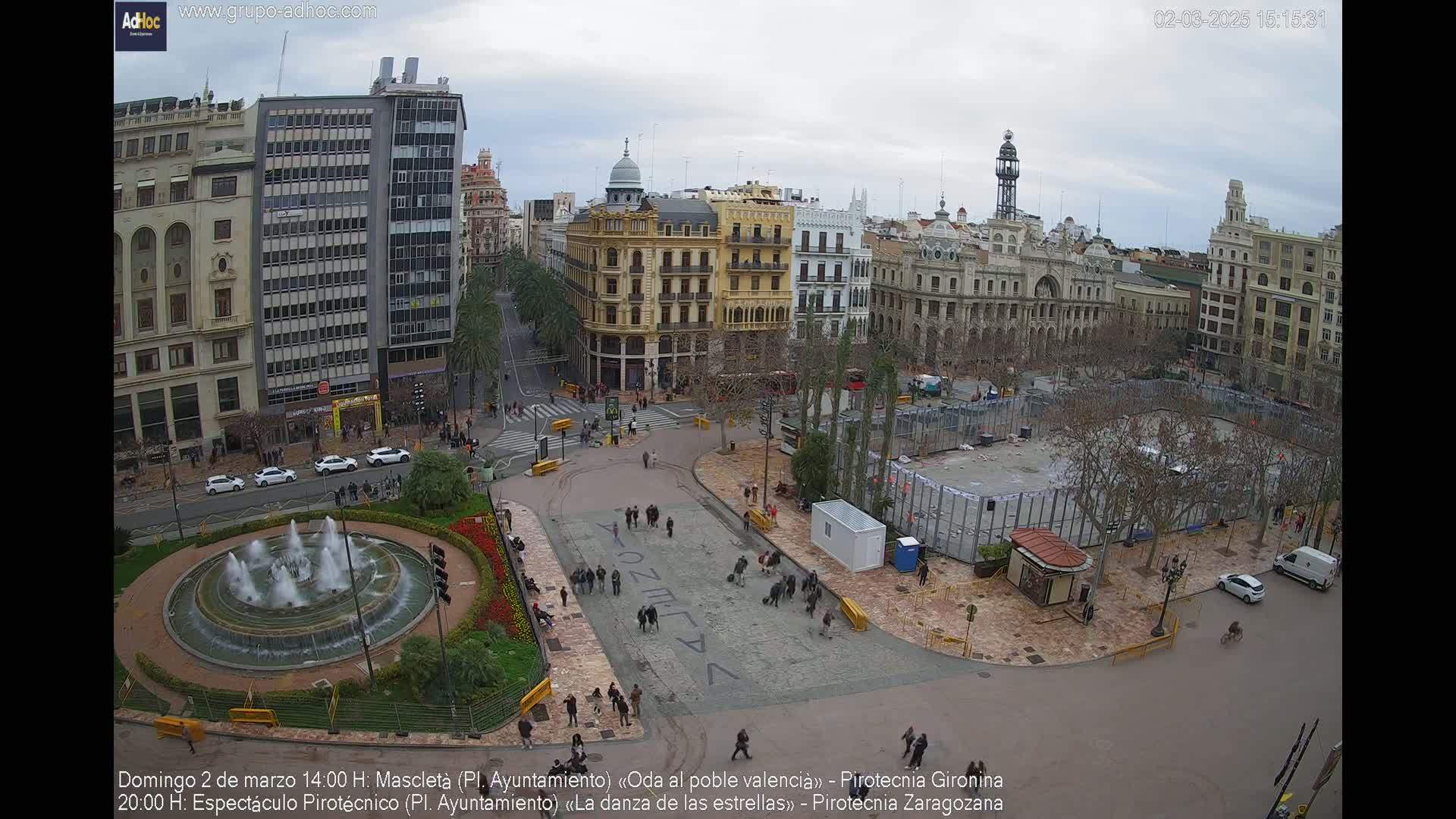 A city square with several buildings surrounding it, a fountain in the foreground, and people scattered throughout under an overcast sky.