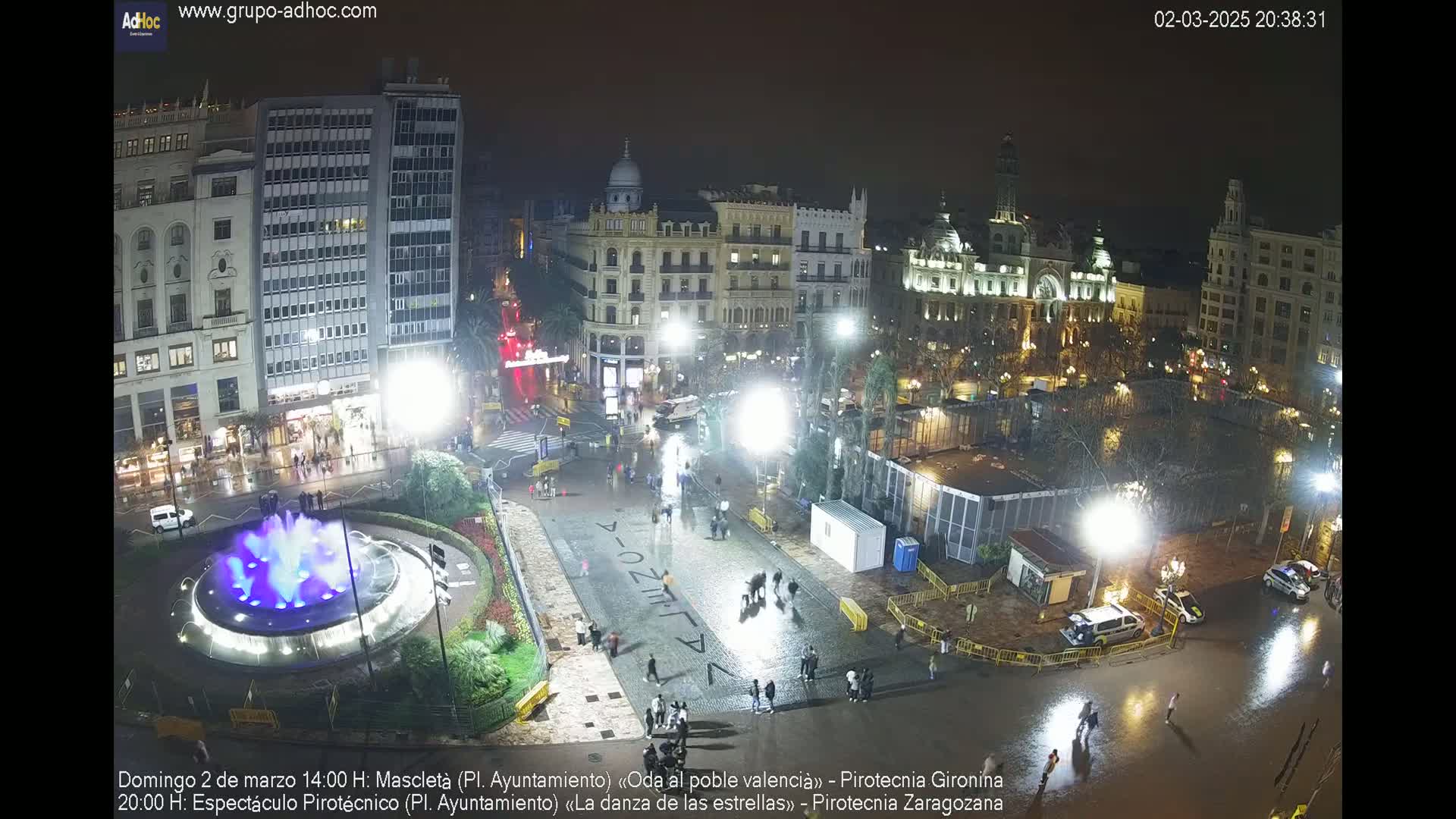 A nighttime, wet city square is illuminated, showing a fountain with blue lights, several people walking around, and buildings in the background.