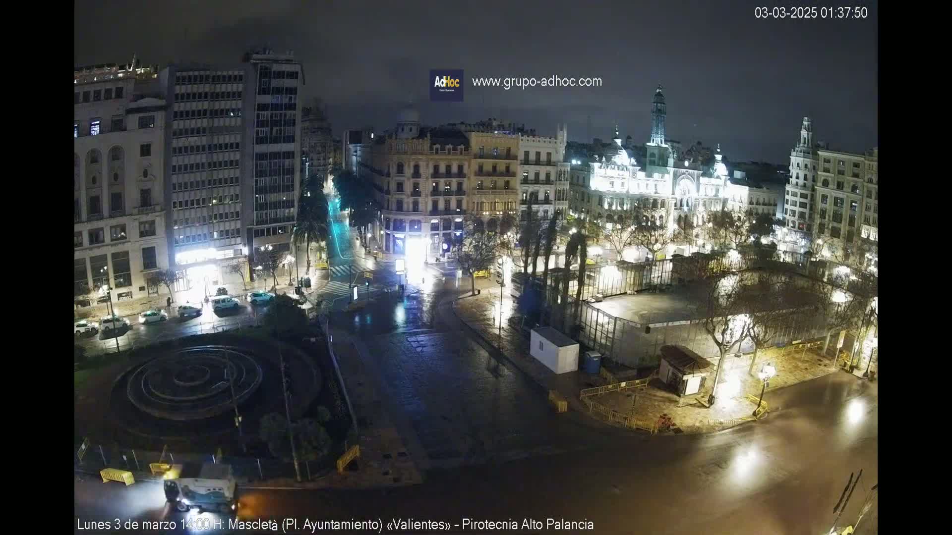 A nighttime, wet city square is seen from above, showing buildings and a circular fountain, with few vehicles and people visible.