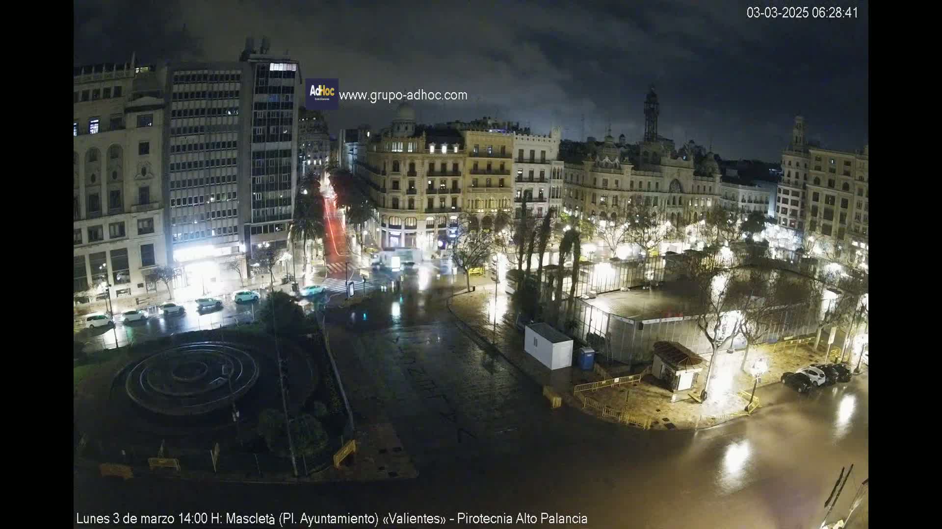 A rain-soaked city square at night, illuminated by streetlights, showing several buildings and a few cars driving on wet roads.
