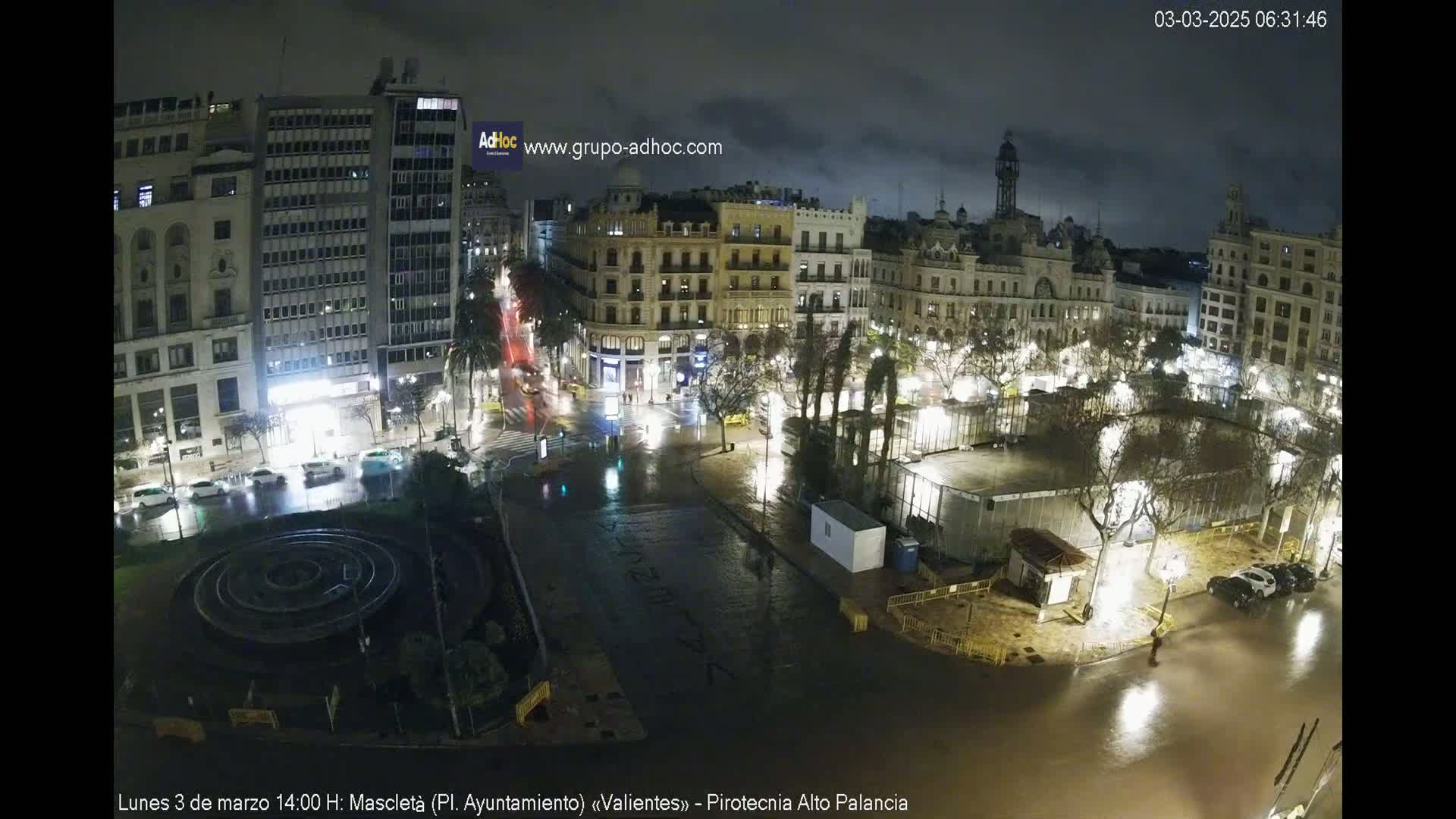 A nighttime city square with buildings surrounding it is flooded with rain, with several cars parked along the streets and some pedestrians visible.