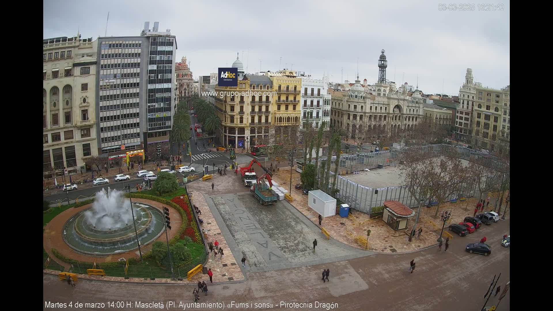 A city square under an overcast sky, features a fountain, construction equipment, and several buildings surrounding it.