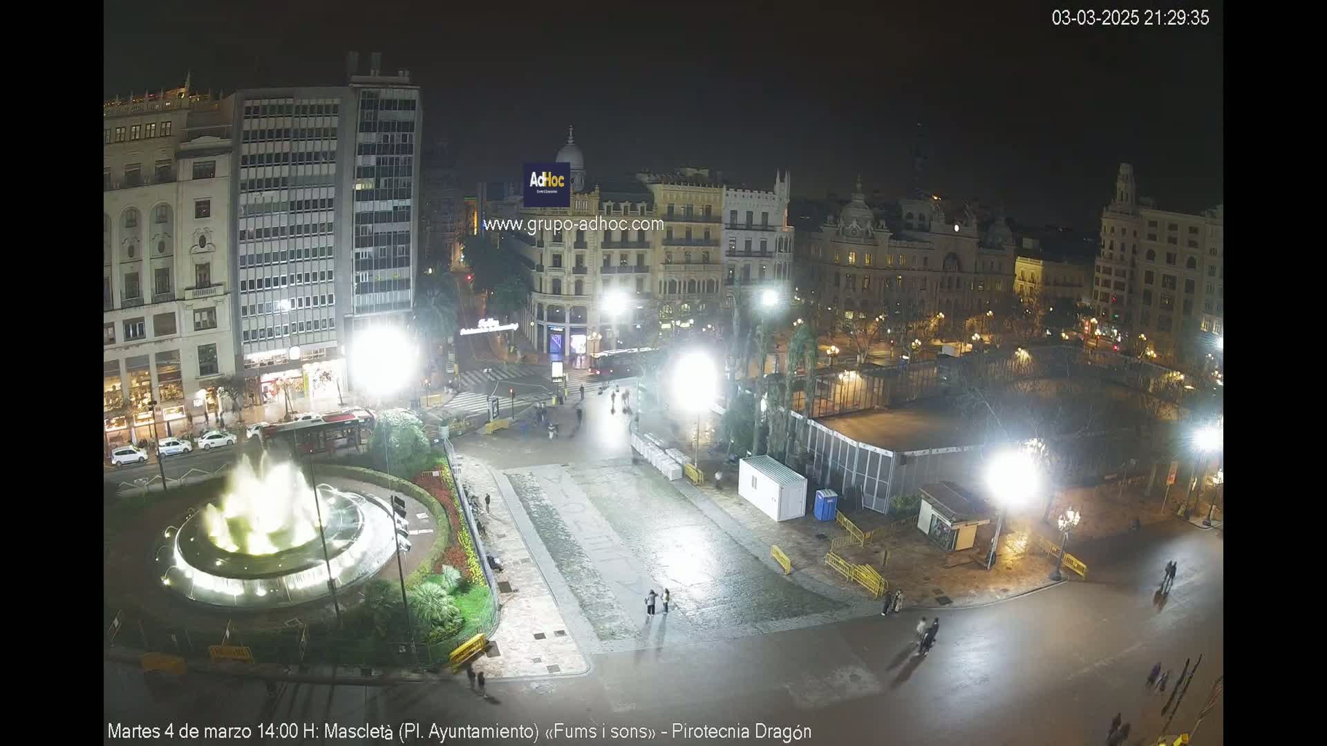 A nighttime, brightly lit city square features a fountain, buildings, and sparsely populated streets; the ground appears wet.