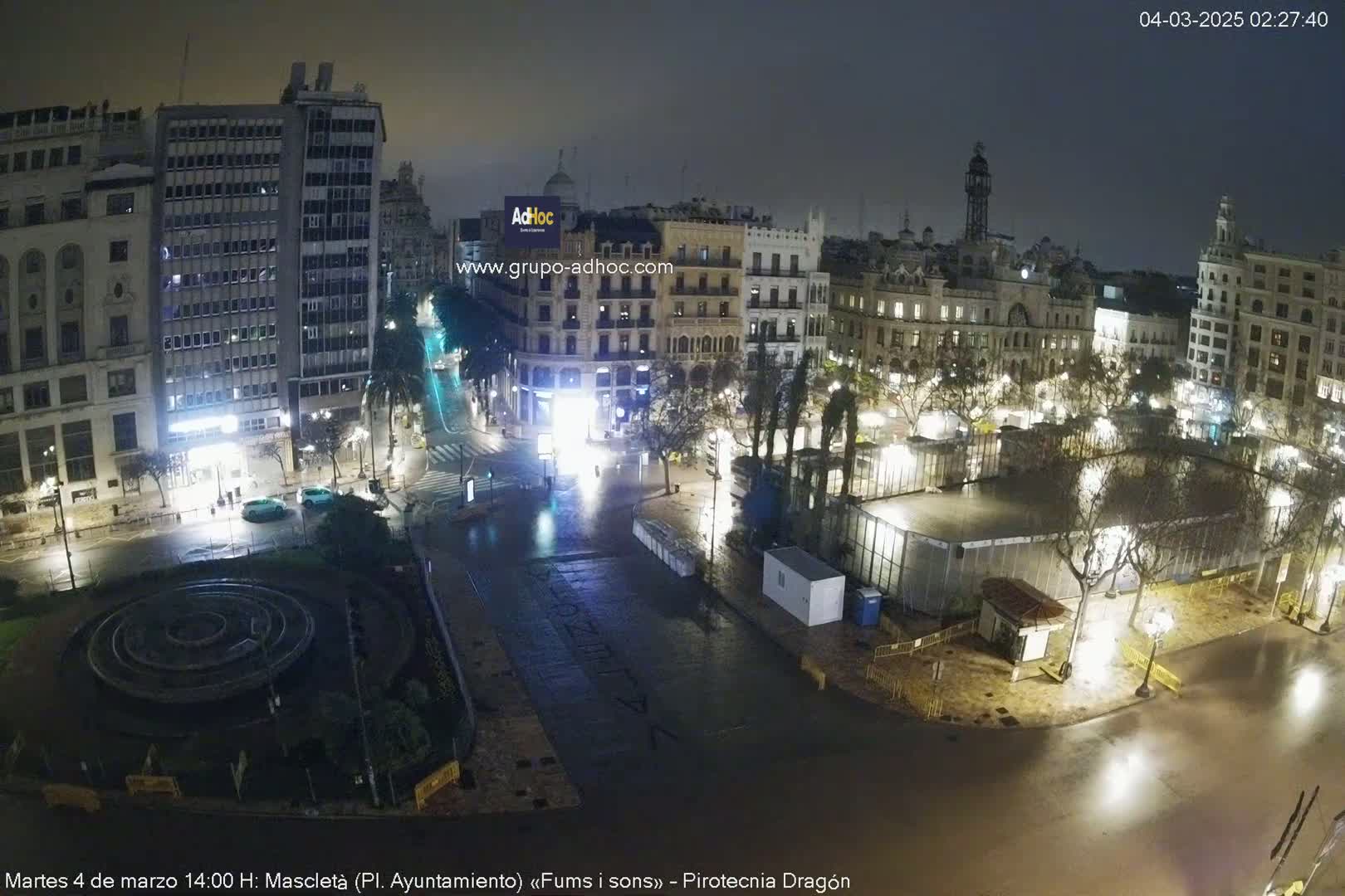A nighttime, wet city square scene shows several buildings, a circular fountain, and temporary structures under dim lighting.