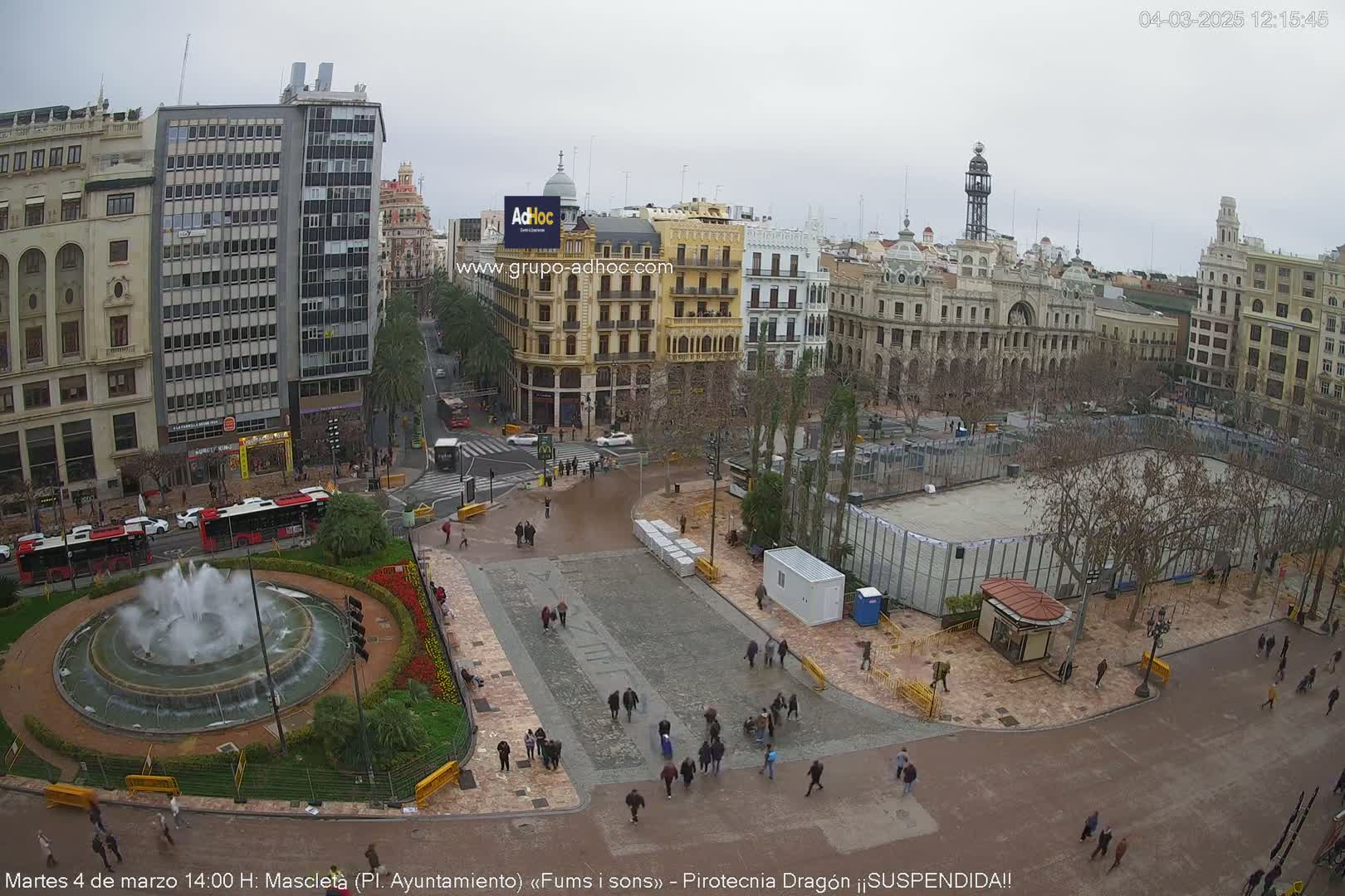 A city square with a fountain, sparsely populated by pedestrians, under an overcast sky;  bare trees and several buildings surround the square.