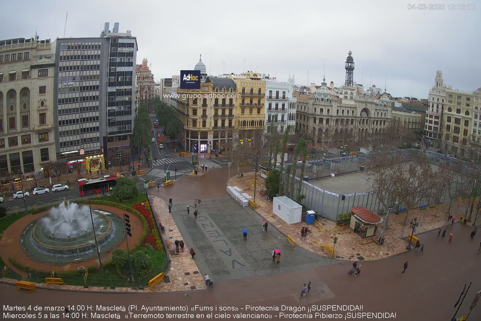 A city square with a fountain, buildings, and people walking around on a cloudy day.