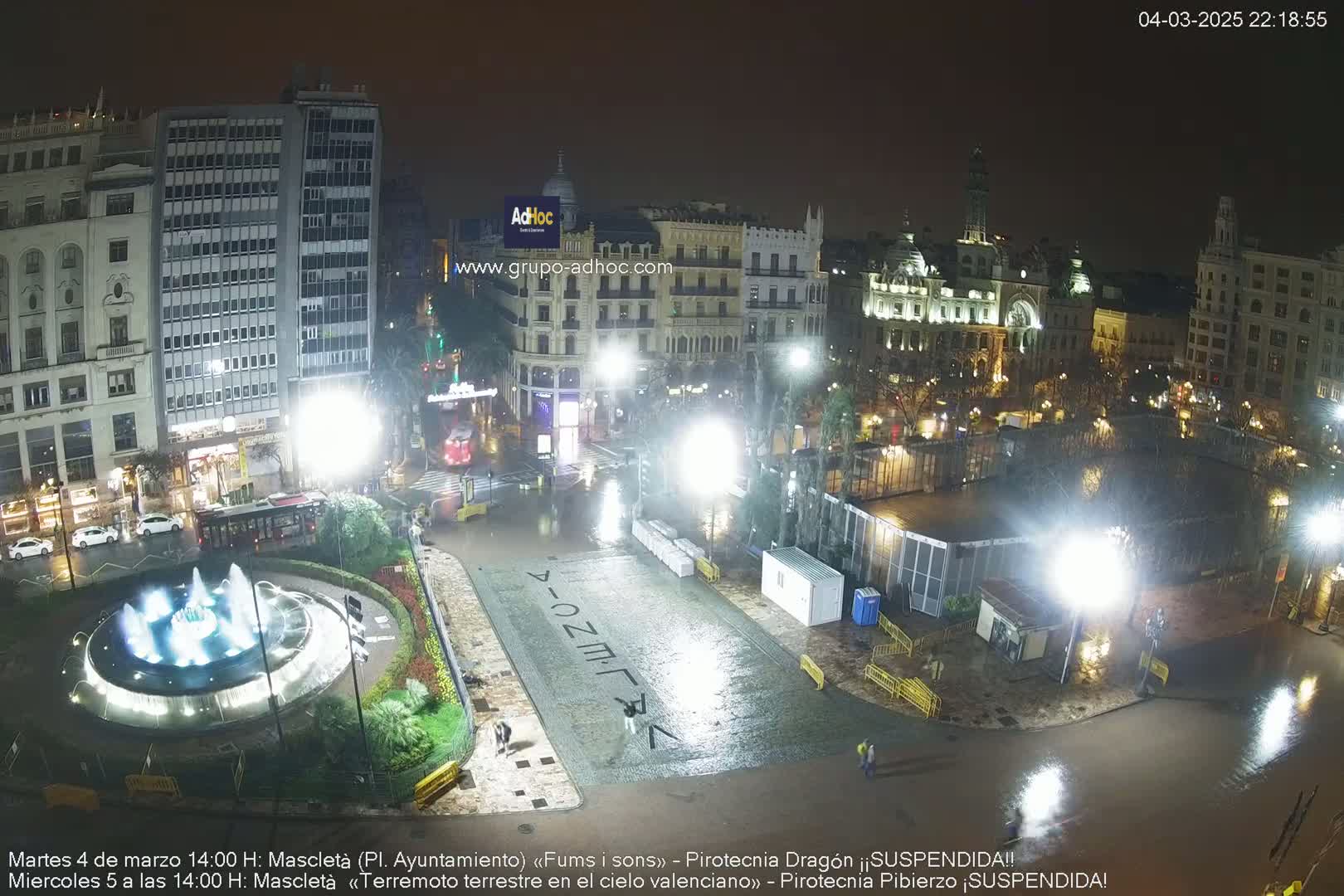 A nighttime, rain-soaked city square features a lit-up fountain, buildings, and temporary structures under bright lights.