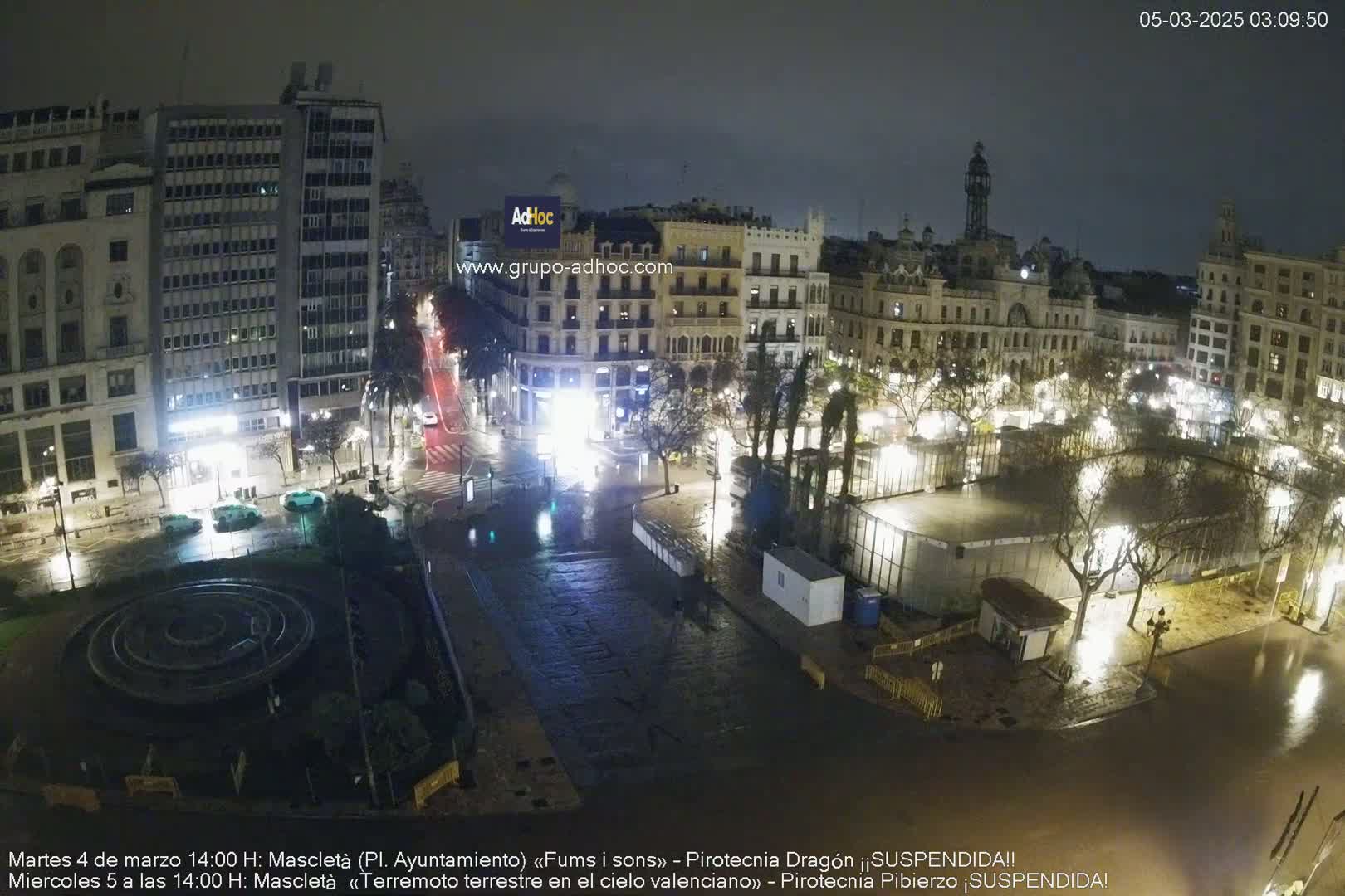 A nighttime, rain-slicked city square with buildings illuminated, a fountain, and some construction in the center.